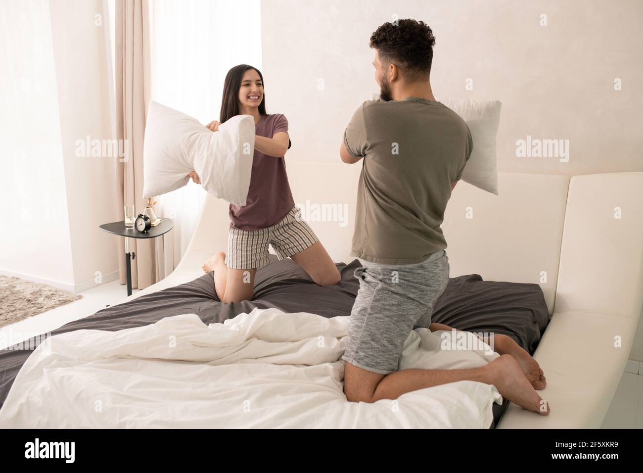 Jeune couple joyeux et amusant exprimant la joie tout en luttant avec des oreillers sur grand lit double le week-end matin après la nuit et petit déjeuner Banque D'Images