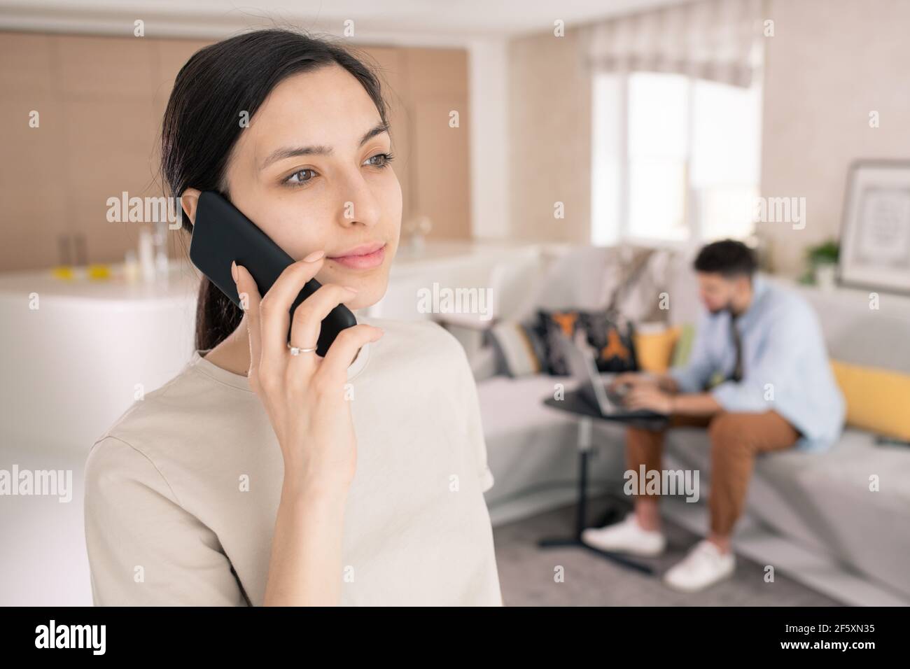 Jeune femme magnifique en t-shirt blanc tenant un smartphone à l'oreille tout en appelant son ami ou collègue contre l'homme à l'aide d'un ordinateur portable sur le canapé Banque D'Images