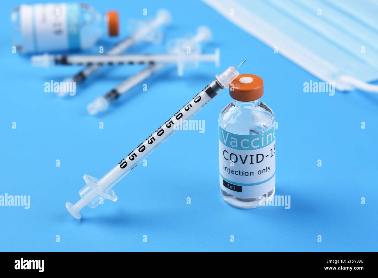 Un flacon de vaccin Covid-19 avec une seringue penchée sur le flacon avec plus de seringues, un flacon et un masque chirurgical en arrière-plan. Banque D'Images