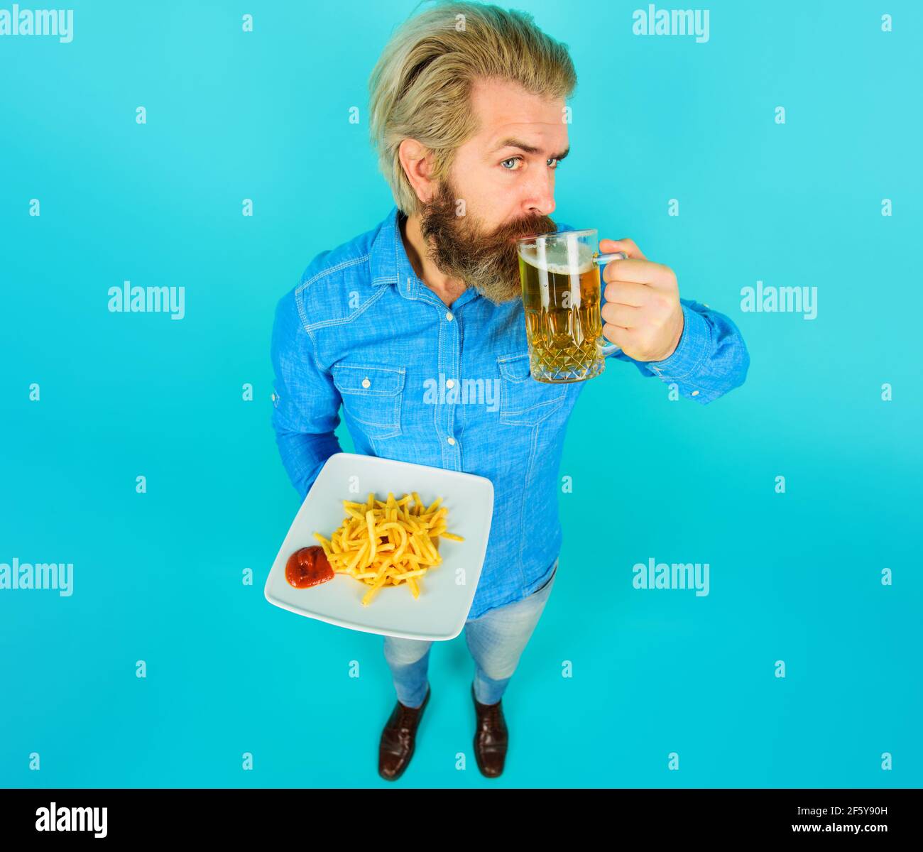 Homme aux frites, ketchup et verre de bière. Un gars barbu avec des pommes de terre frites. Restauration rapide, mauvaise alimentation, malbouffe. Banque D'Images