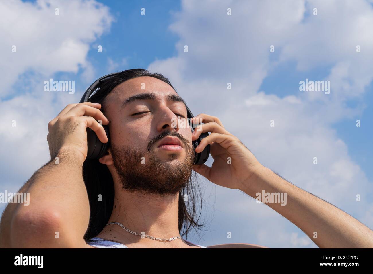 jeune homme barbu avec casque sans fil avec le ciel en arrière-plan, réfléchissant sur la vie, méditant. avec les yeux fermés. son surround. Banque D'Images