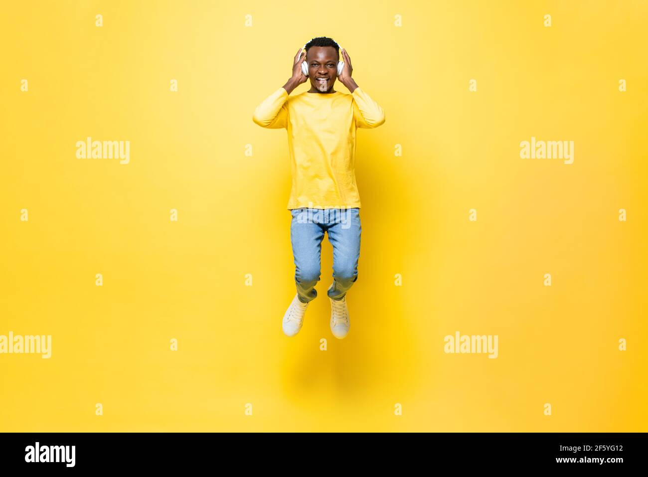 Un jeune homme africain heureux portant un casque à l'écoute de musique et saut sur fond de studio isolé jaune Banque D'Images