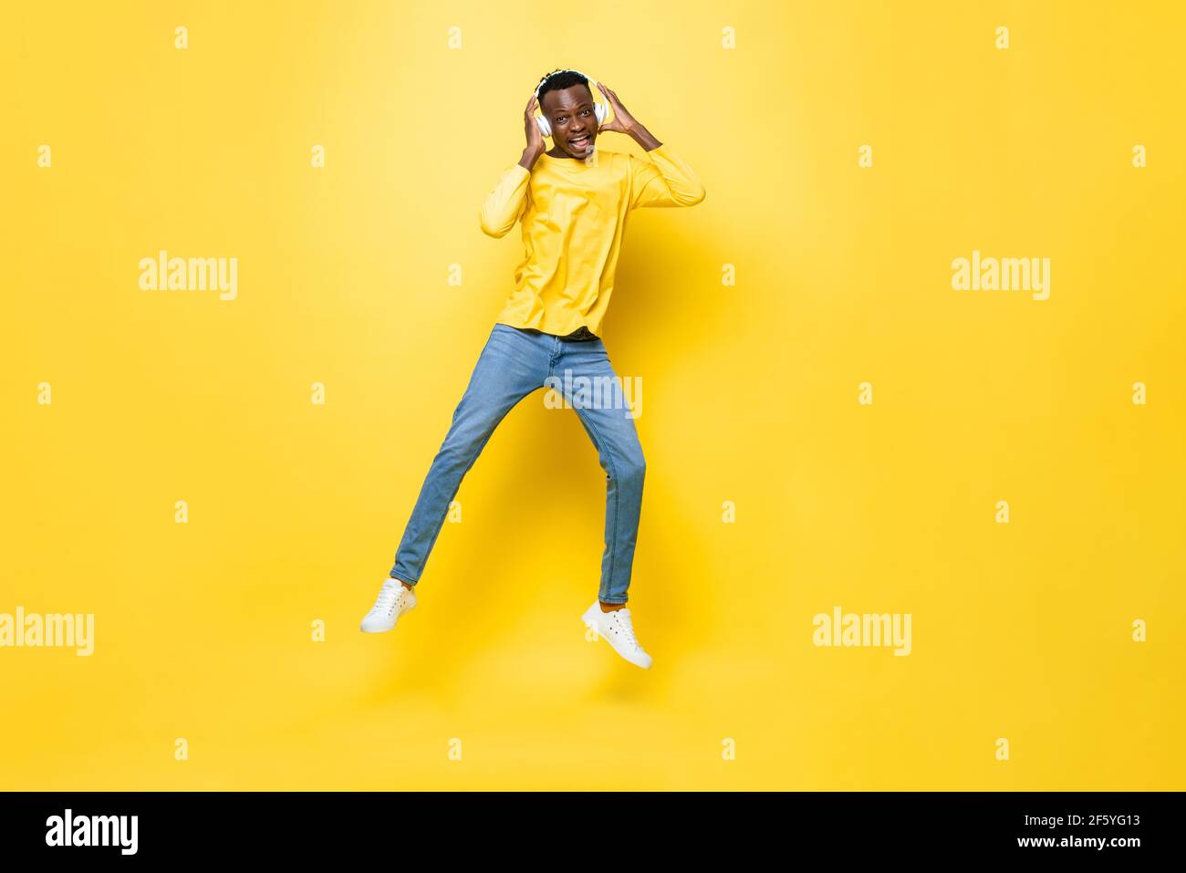 Un jeune homme africain heureux portant un casque à l'écoute de musique et saut sur fond de studio isolé jaune Banque D'Images