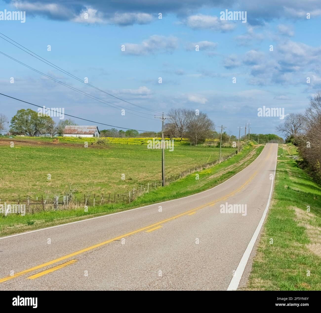 Route de campagne vide avec un pâturage clôturé et une grange sur la gauche sur une partie nuageux tôt le jour de printemps. Banque D'Images