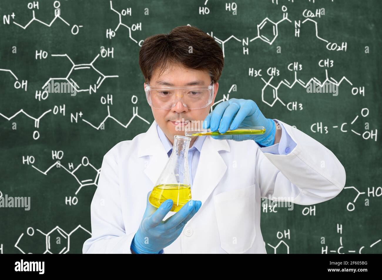 Un scientifique asiatique de milieu d'année portant des gants et tenant un conteneur expérimental devant un tableau noir avec une formule écrite dessus. Banque D'Images