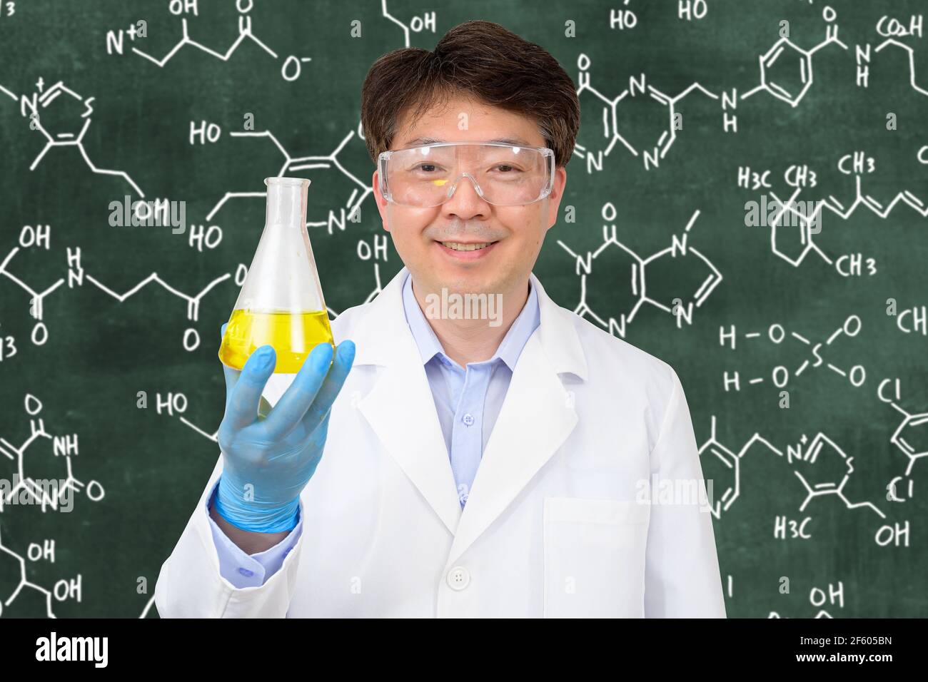 Un scientifique asiatique de milieu d'année portant des gants et tenant un conteneur expérimental devant un tableau noir avec une formule écrite dessus. Banque D'Images