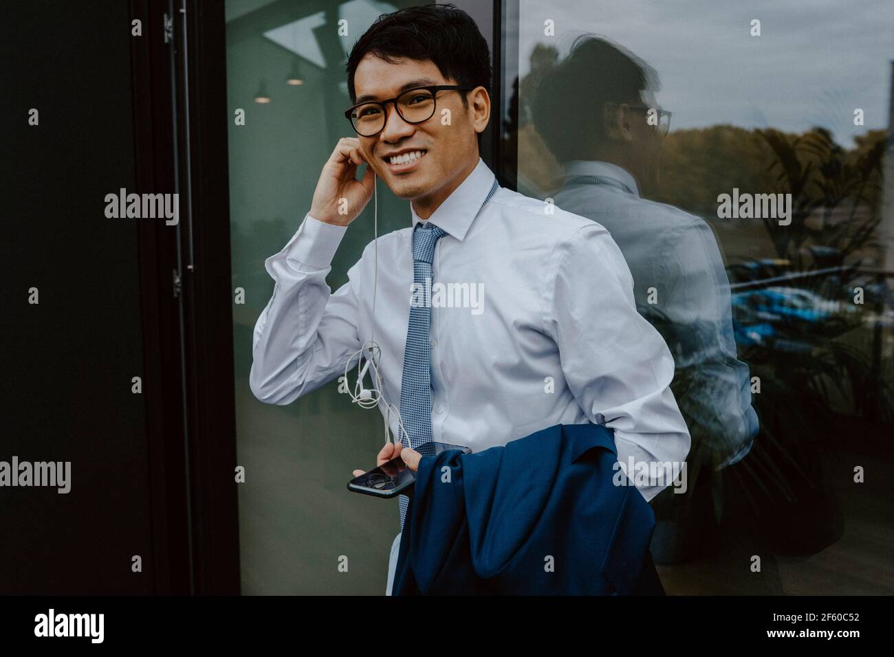 Portrait d'un homme d'affaires souriant écoutant de la musique à l'aide d'écouteurs tout en étant debout contre la fenêtre Banque D'Images