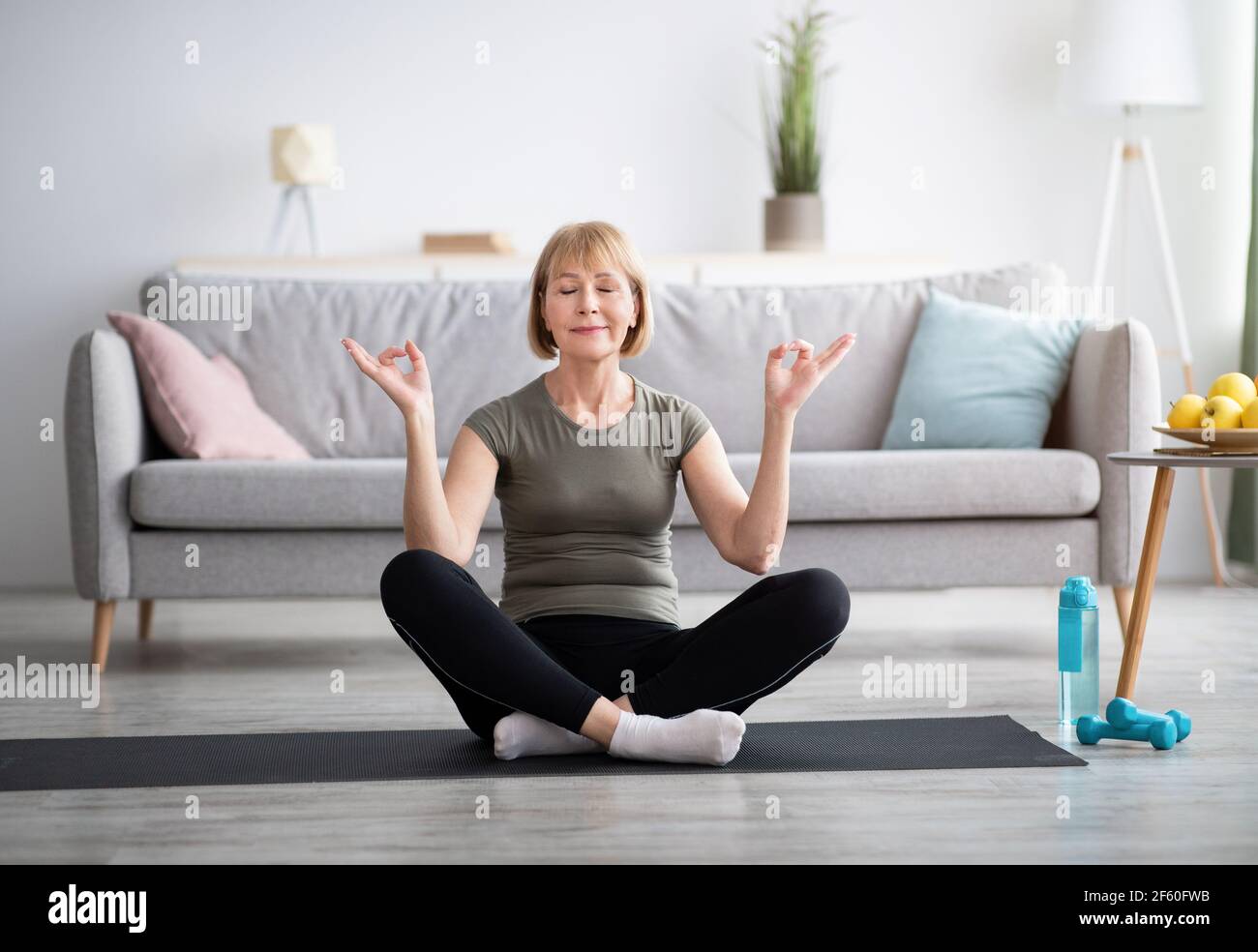 Pleine longueur de femme mûre méditant avec les yeux fermés, faisant gyan mudra avec ses mains à la maison Banque D'Images