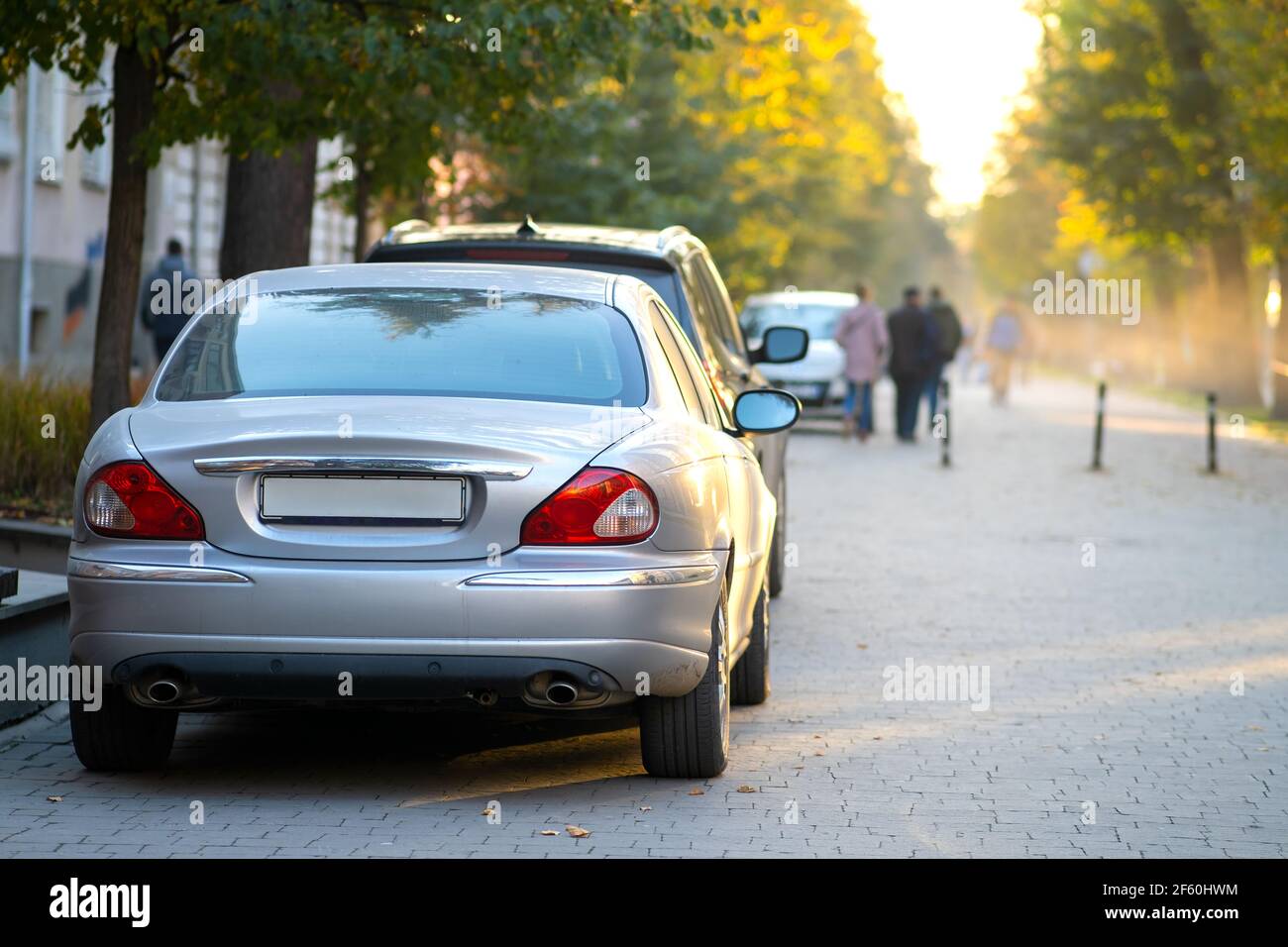 Voitures garées dans une rangée sur le côté de la rue de la ville le jour d'automne lumineux avec des personnes floues marchant sur la zone piétonne. Banque D'Images