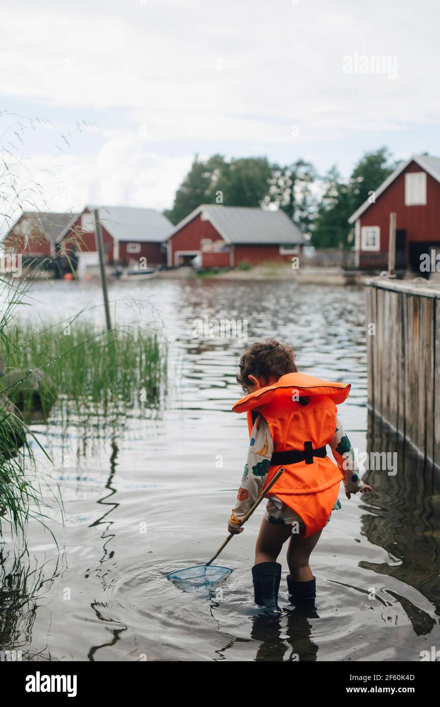 Vue arrière du garçon portant un gilet de sauvetage debout lac avec canne à pêche en été Banque D'Images