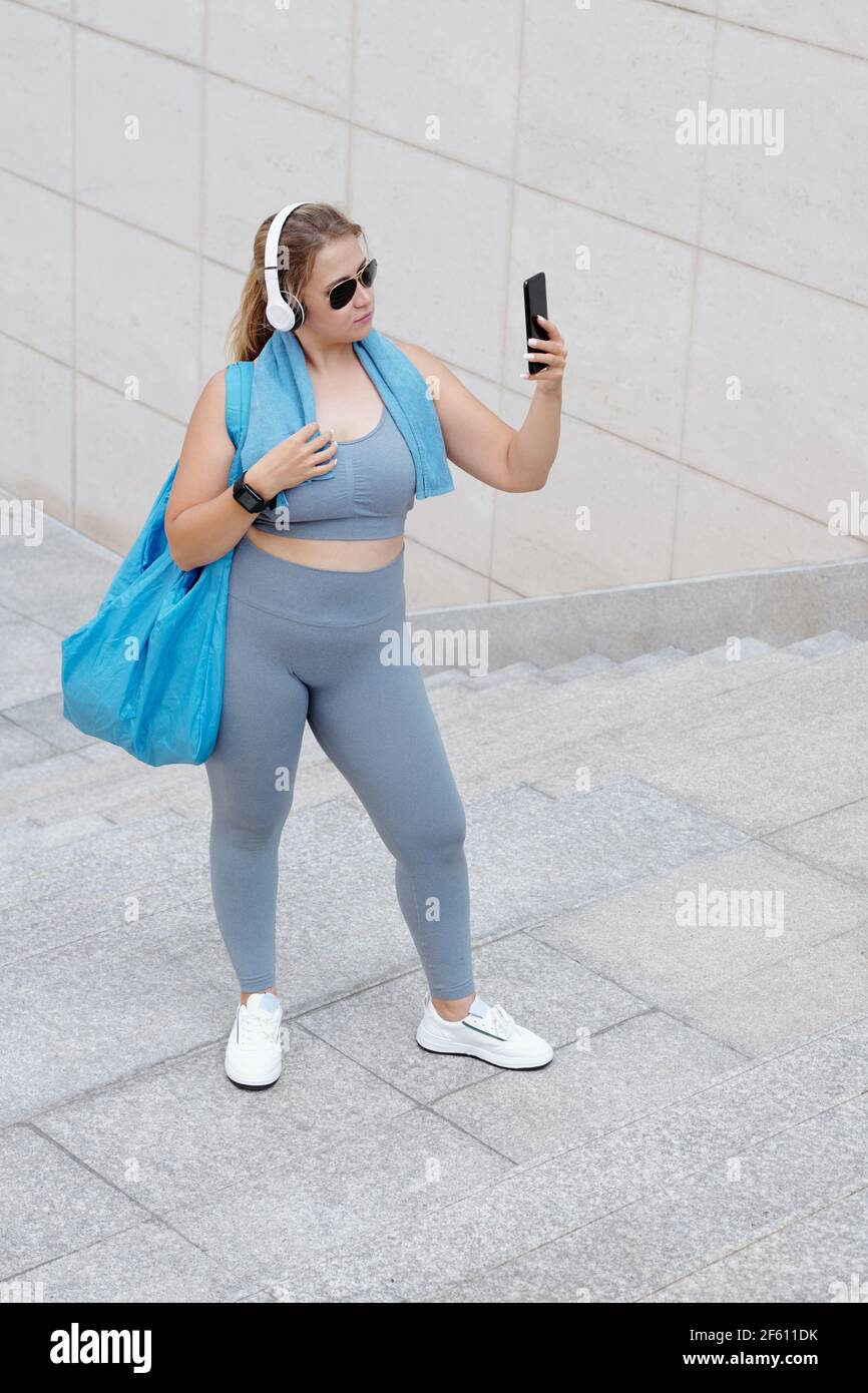 Jolie femme de taille plus avec un sac de gym posé pour selfie après l'entraînement en lunettes de soleil et écouteurs Banque D'Images