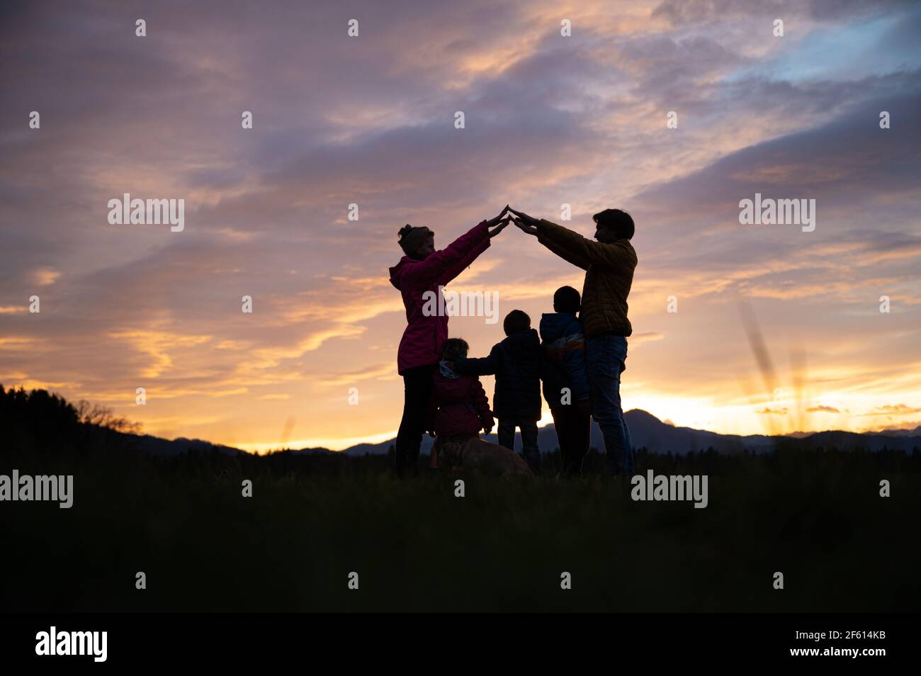Silhouette d'une famille à l'extérieur sous un beau ciel nocturne avec des parents qui font une maison avec leurs bras sur leurs trois enfants et un chien. Banque D'Images