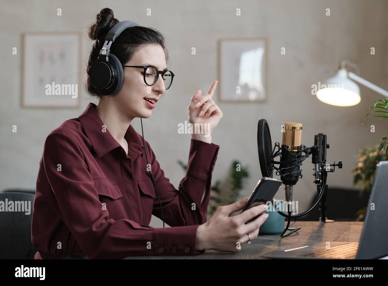 Jeune DJ radio dans un casque parlant en ligne à l'aide de son mobile téléphone à la table en studio Banque D'Images