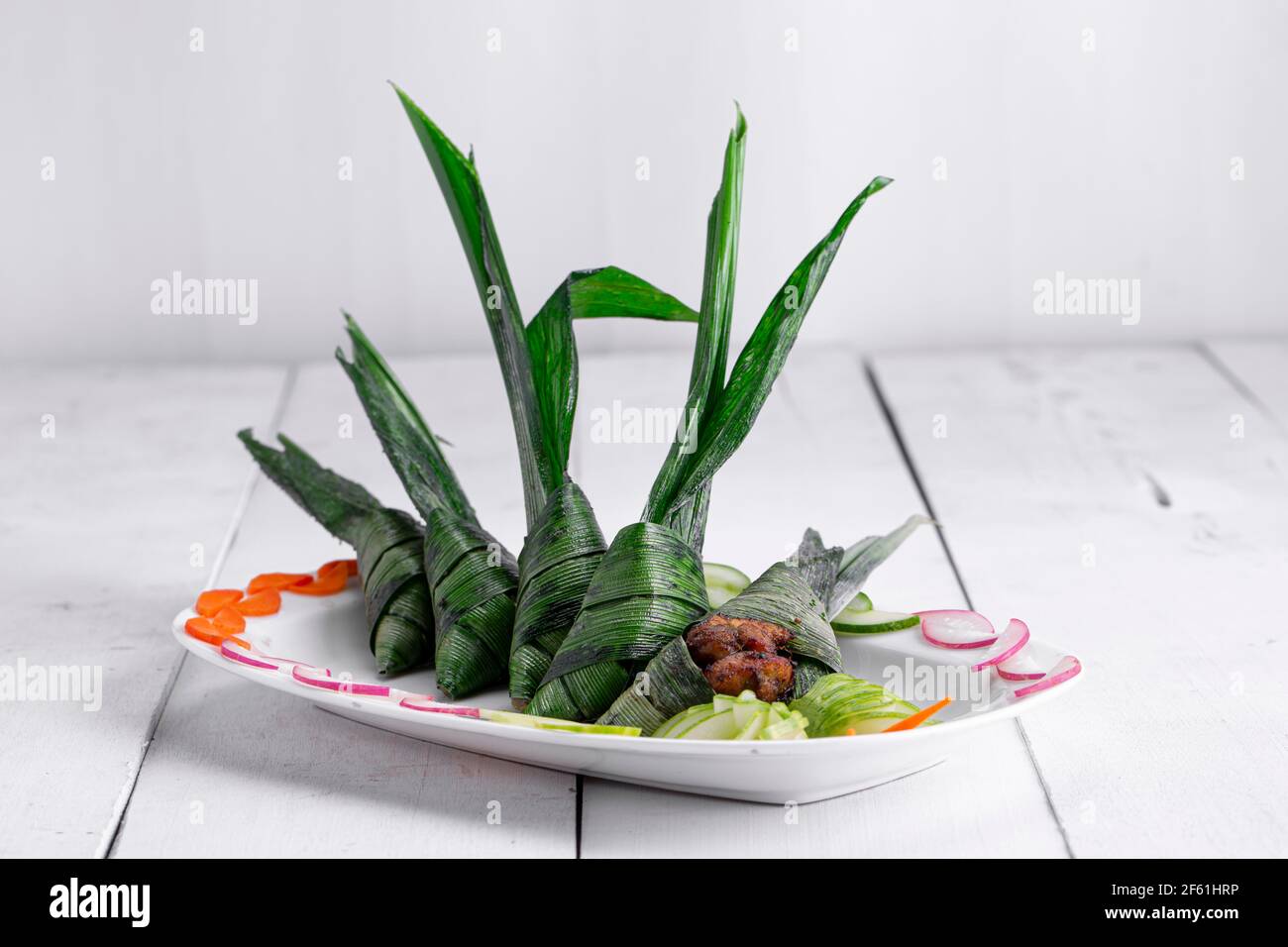 Poulet enveloppé de feuilles de palmier placé magnifiquement dans une plaque en céramique blanche avec fond blanc texturé et sauce placée à proximité. Banque D'Images