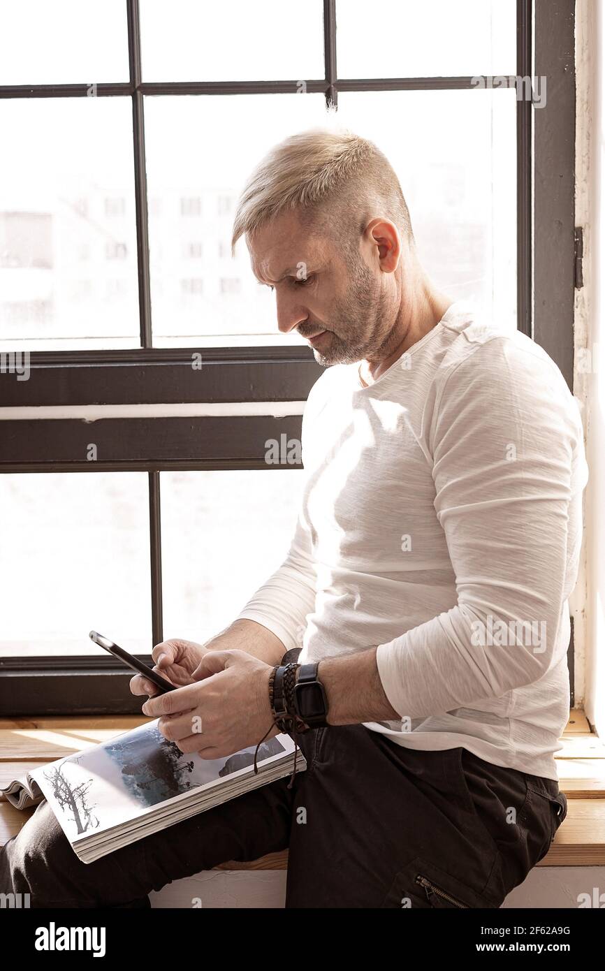 Beau homme concentré dans une chemise blanche regarde son téléphone mobile assis sur un rebord de fenêtre. Origine ethnique caucasienne, jour ensoleillé, travail et affaires, Banque D'Images