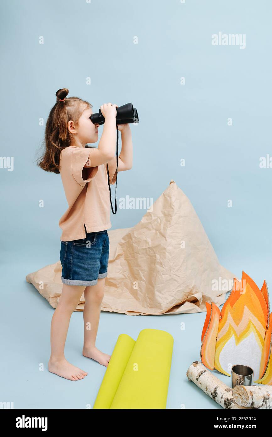 Petite fille curieuse debout à côté d'un faux feu de camp et d'une tente en papier, regardant à travers des jumelles. Sur fond bleu Banque D'Images