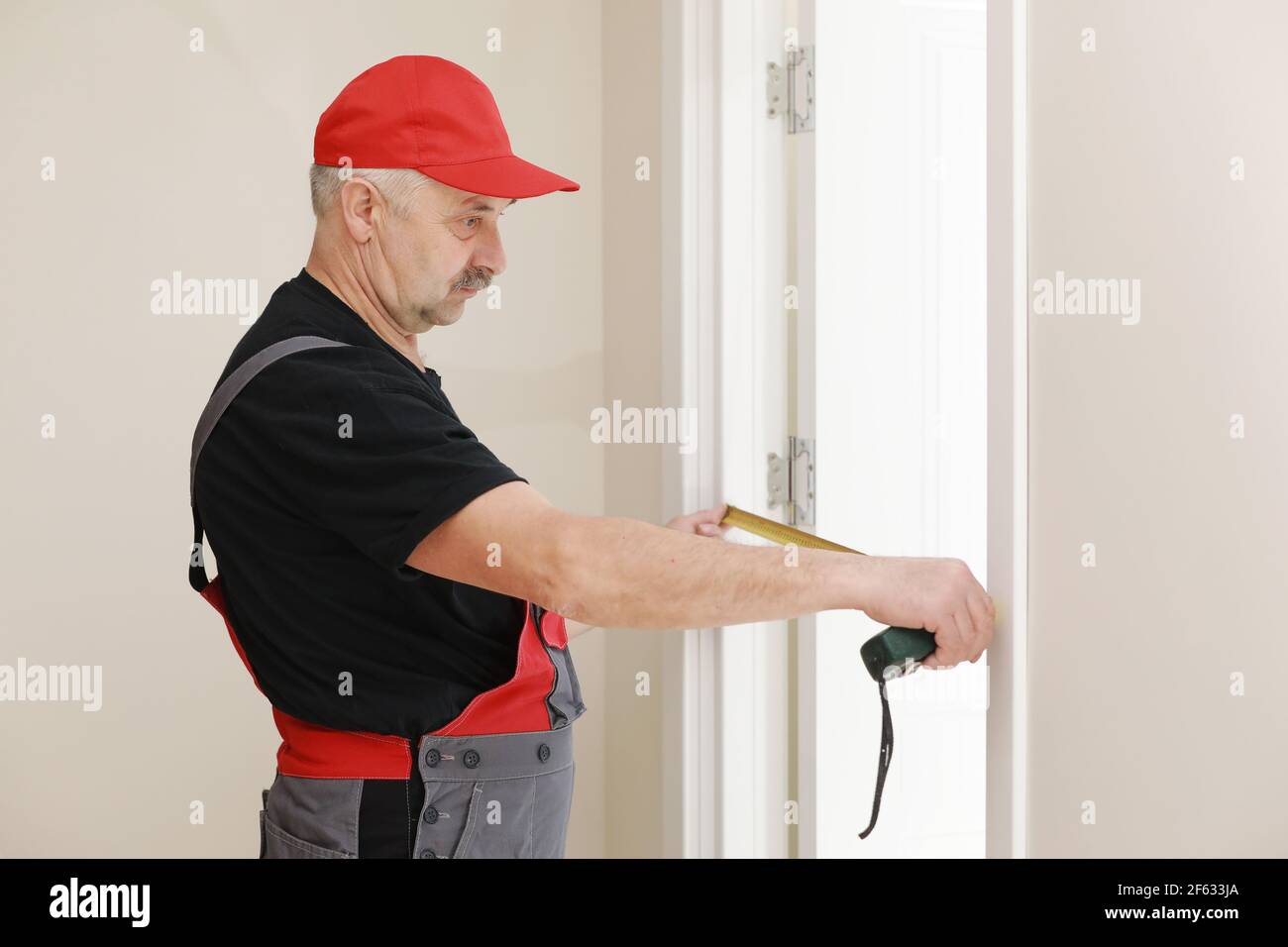 Ouvrier en casquette rouge et combinaison de travail avec le ruban de  mesure. L'homme installe les portes. Mesurer le ruban dans les mains.  Travaux de réparation. Entretien dans les appartements Photo Stock -