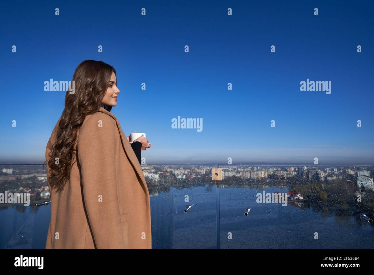 Belle jeune femme buvant du café le matin tout en se tenant sur le balcon. Une dame agréable jouissant d'une vue magnifique sur la grande ville. Concept de détente. Banque D'Images