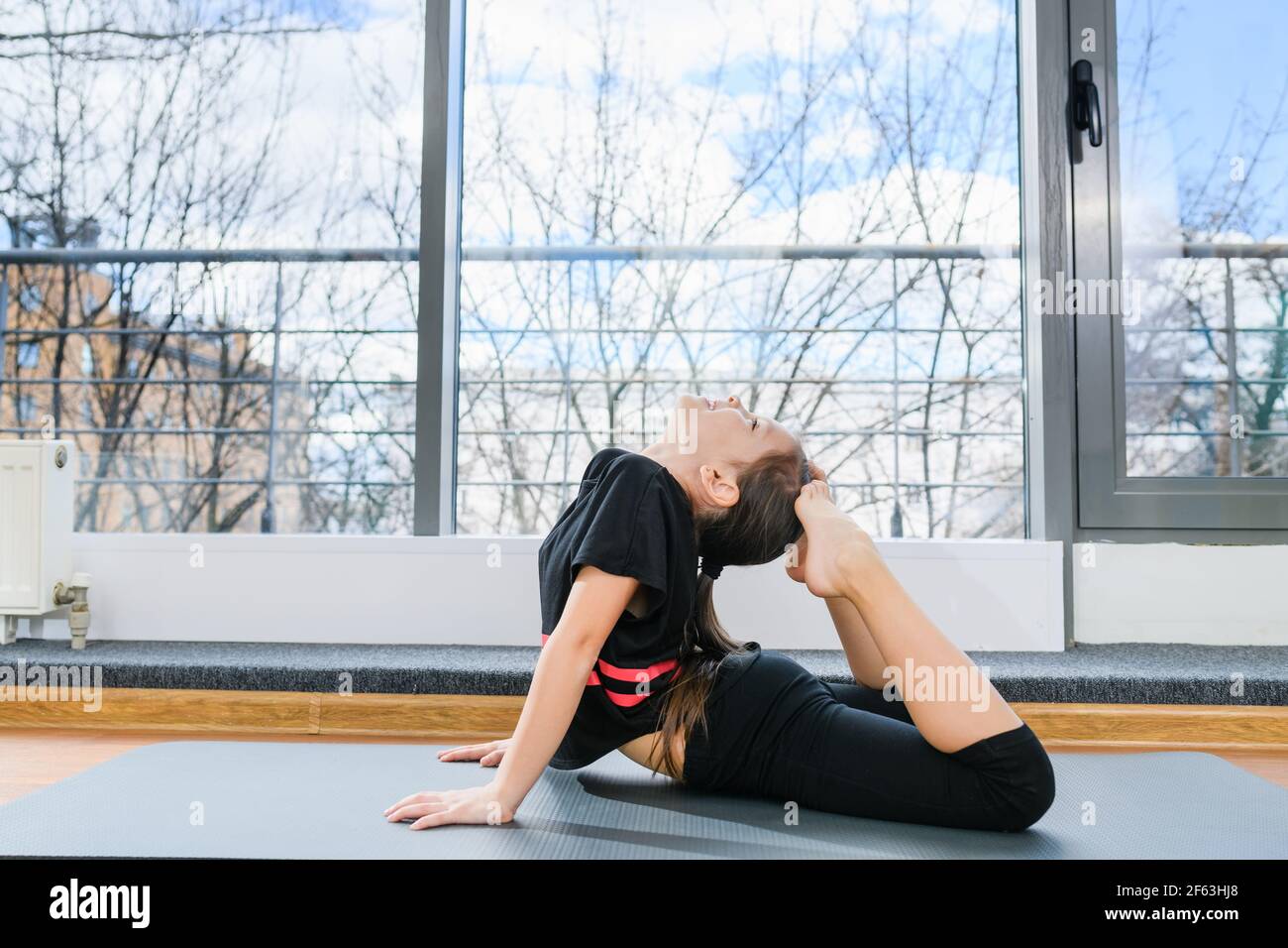 Préteen flexible fille couché sur le ventre et toucher la tête par les pieds dans la salle de lumière avec fenêtre panoramique, gymnastique pour les enfants Banque D'Images
