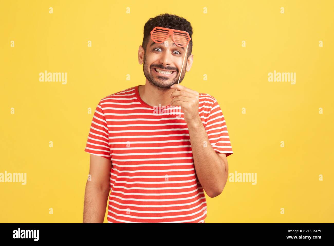 Un homme enthousiaste avec une barbe en t-shirt rayé tenant un masque de fête sur le bâton et regardant l'appareil photo avec un sourire crasseux, en s'amusant. Prise de vue en studio Banque D'Images