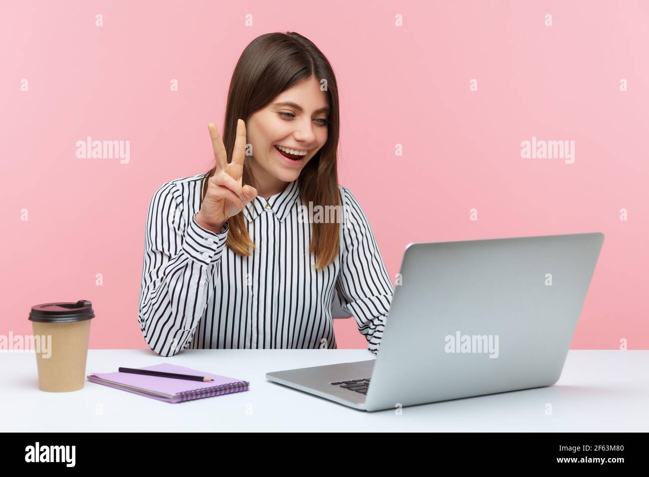 Une employée de bureau positive réussie montrant le geste de victoire et souriant regardant dans l'écran d'ordinateur portable, parlant appel vidéo, conférence en ligne. Intérieur Banque D'Images