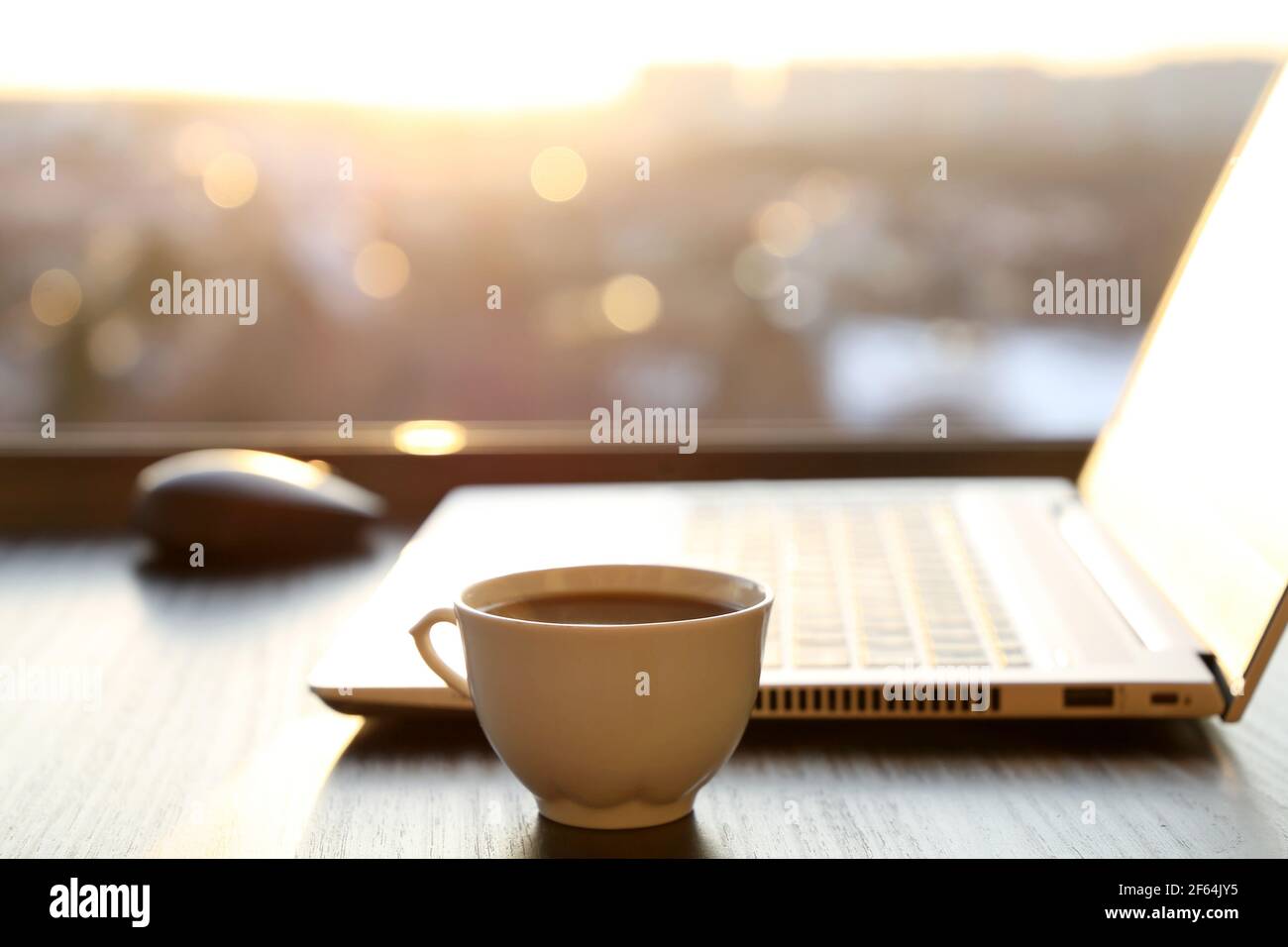 Espace de travail confortable dans le bureau à la maison avec une tasse de café chaude et un ordinateur portable sur la table contre la fenêtre au coucher du soleil. Concept de travail à distance et de gain en ligne Banque D'Images