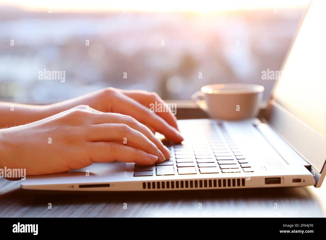 Femme assise au clavier de l'ordinateur portable près de la fenêtre au coucher du soleil, les mains femelles se rapprochent de la lumière du soleil. Espace de travail confortable avec tasse à café Banque D'Images