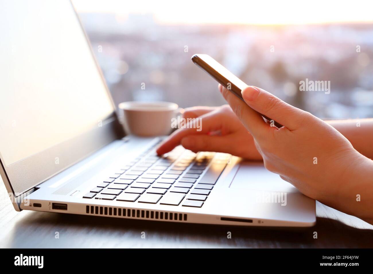 Femme avec un smartphone assis au clavier de l'ordinateur portable près de la fenêtre au coucher du soleil. Espace de travail confortable, concept de communication en ligne, bureau ou travail à domicile Banque D'Images