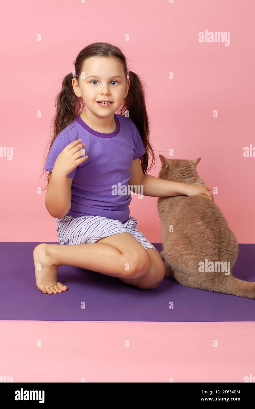 Portrait intégral d'une fille aux queues de cheval dans un t-shirt violet et un short tenant un chat rouge sur un tapis de sport violet, isolé sur un fond rose Banque D'Images