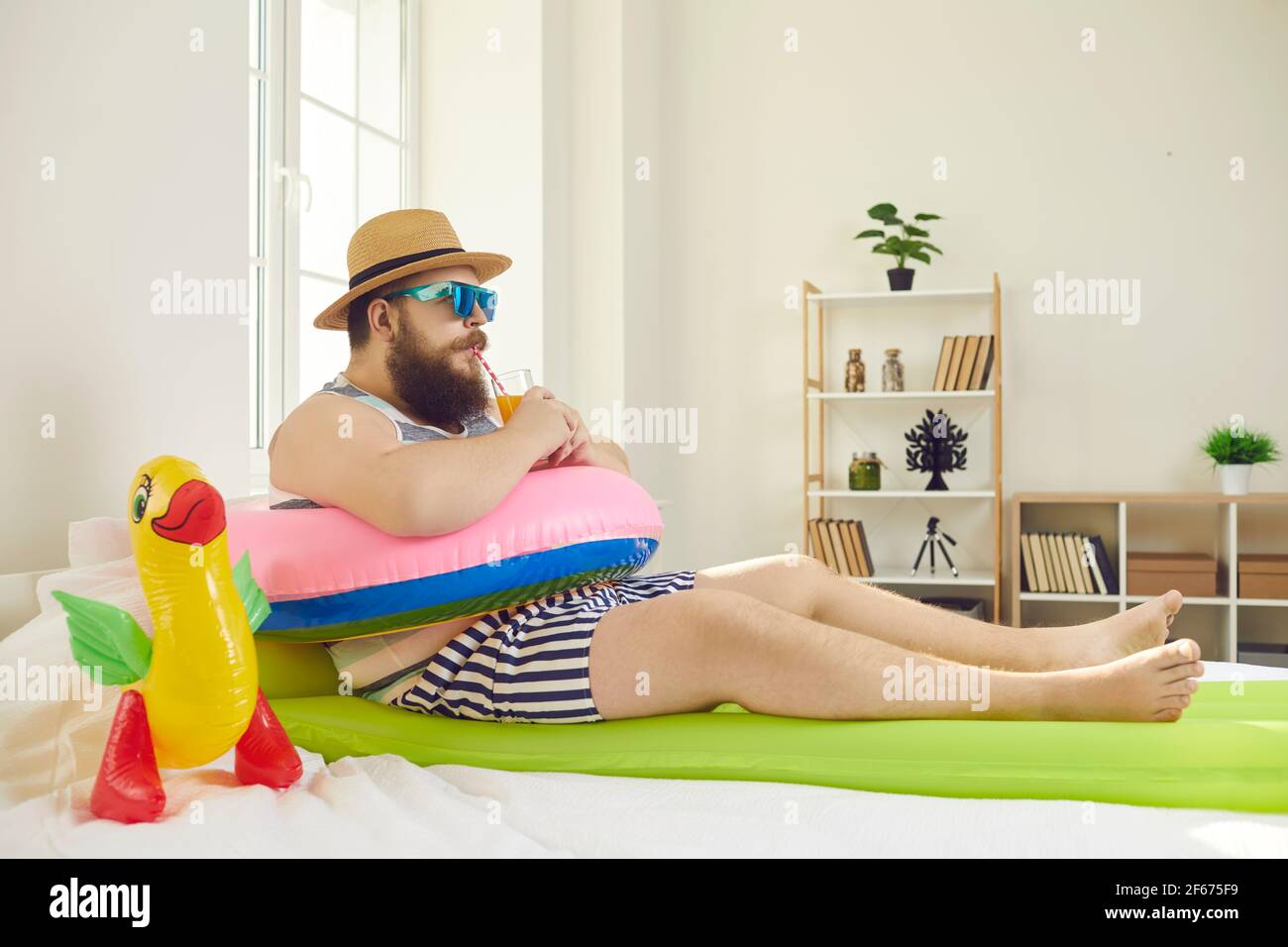 Drôle d'homme dans des lunettes de soleil et chapeau de soleil boire jus assis sur la plage, il y a un anneau de natation à la maison Banque D'Images