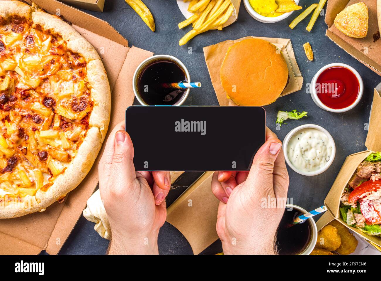 Livraison fastfood commande de nourriture en ligne concept. Grand assortiment de plats à emporter, mains de homme avec smartphone en vue panoramique Banque D'Images