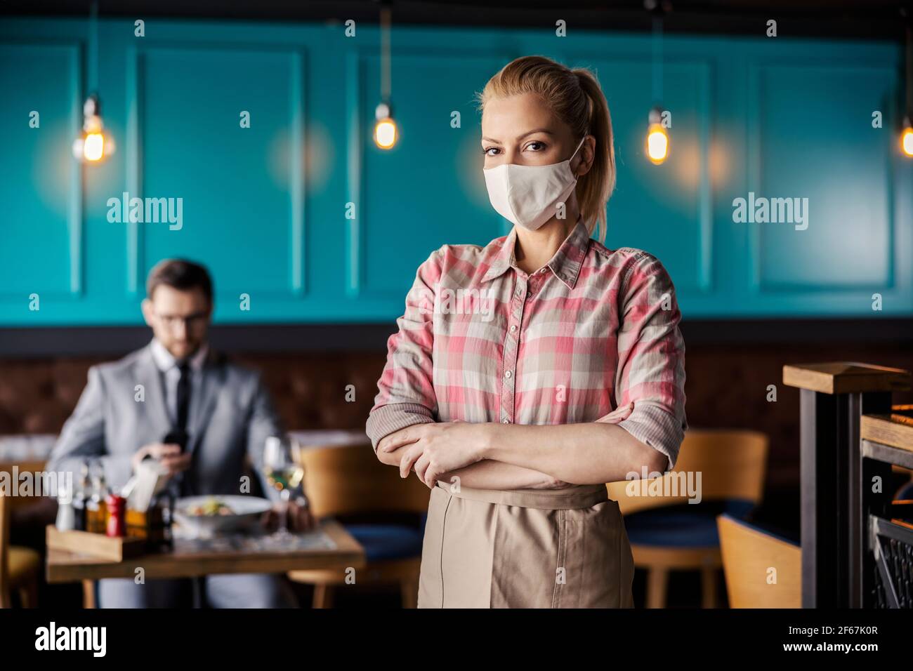 Portrait d'une serveuse debout dans un restaurant intérieur moderne avec ses bras croisés. Une fille blonde dans une chemise à carreaux et un tablier beige porte un prote Banque D'Images