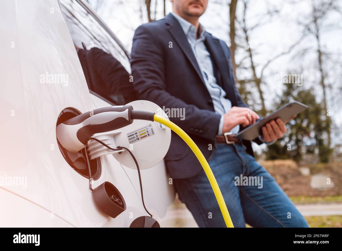 Homme d'affaires debout près d'une voiture électrique en charge ou d'une voiture électrique et en utilisant une tablette dans la rue à la lumière du soleil Banque D'Images