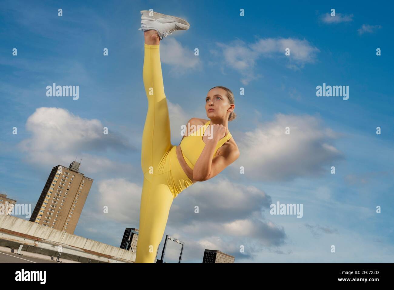 Femme sportive attrayante portant des vêtements d'activité jaunes pratiquant un exercice de coup de pied élevé à l'extérieur dans un cadre urbain. Banque D'Images