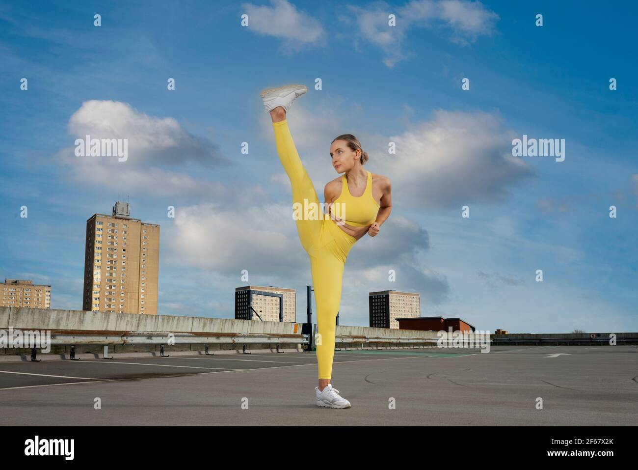 Femme sportive attrayante portant des vêtements d'activité jaunes pratiquant un exercice de coup de pied élevé à l'extérieur dans un cadre urbain. Banque D'Images