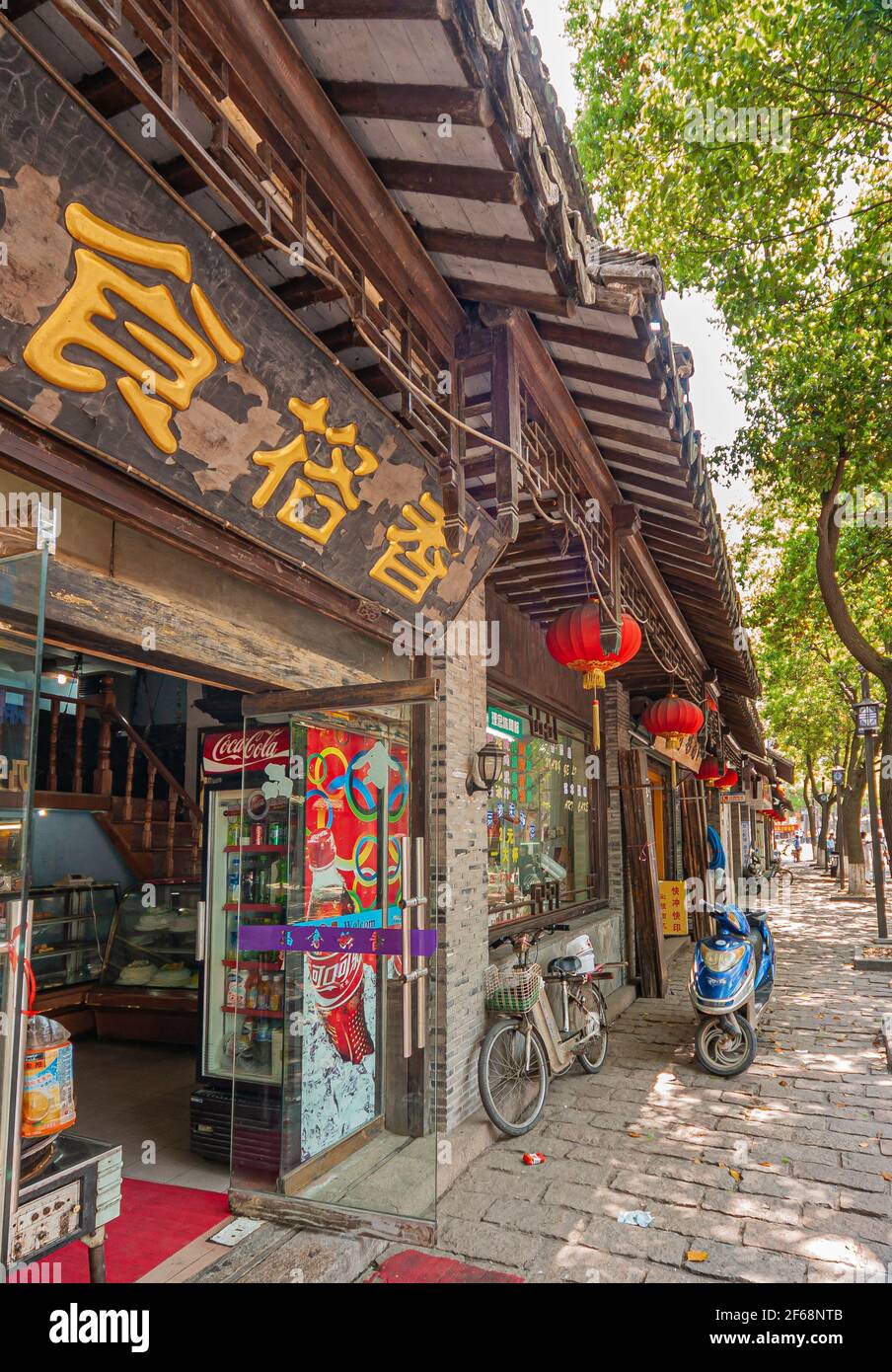 Tongli, Chine - 2 mai 2010 : rangées de petites boutiques avec des avant--toits en bois foncé. Un peu de feuillage vert sur le côté. Lanternes rouges et dorées le long. Scooter bleu a Banque D'Images