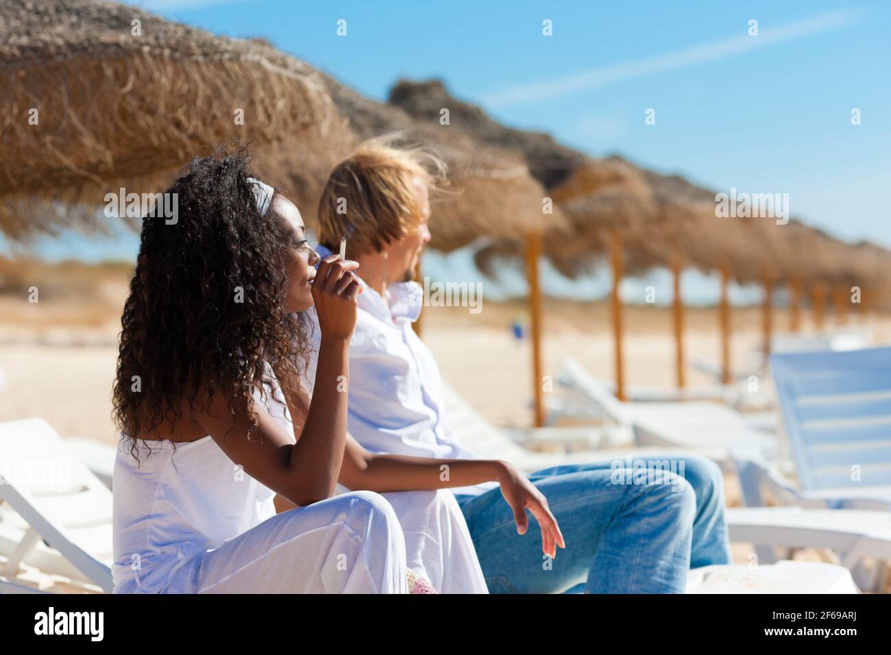 Couple assis dans des chaises longues sur une plage appréciant le soleil fume une cigarette Banque D'Images