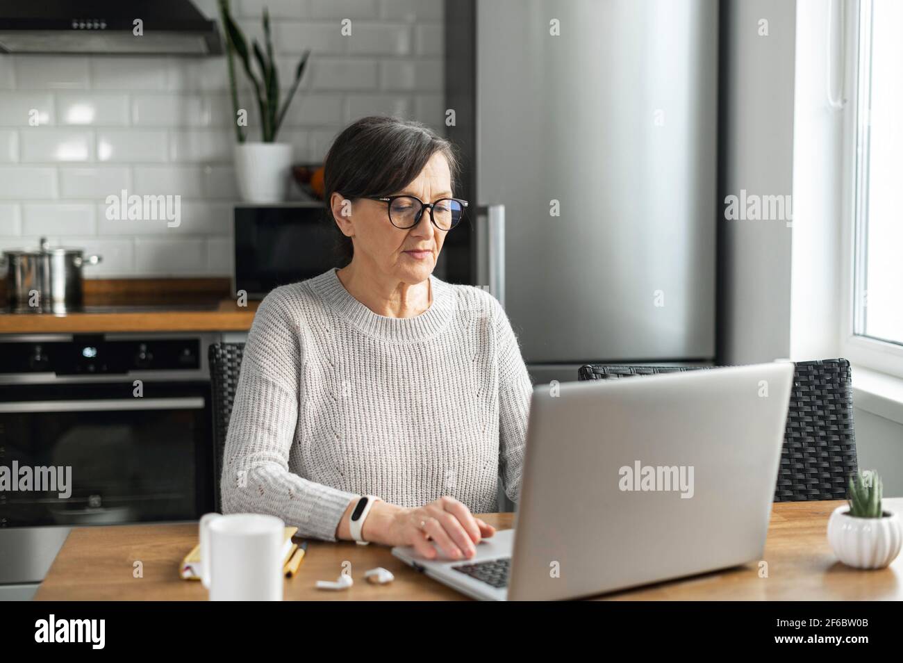 Femme à la retraite utilisant un ordinateur portable pour le travail à distance, femme âgée moderne en lunettes se trouve au bureau de la cuisine et dactylographiant des e-mails, effectue de la correspondance d'affaires à distance Banque D'Images