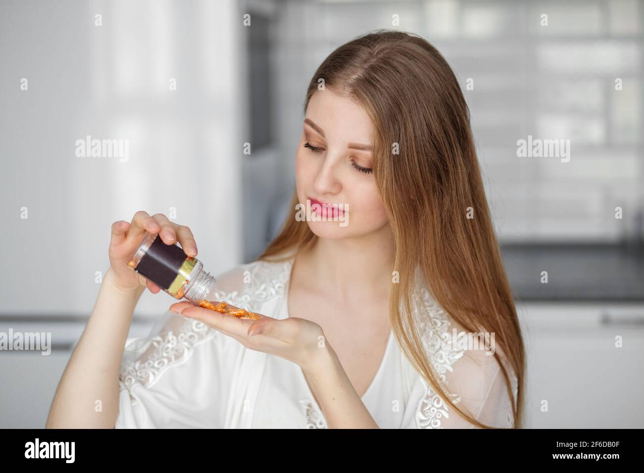 Une belle femme en vêtements blancs montre verser plusieurs comprimés de suppléments alimentaires de vitamines d'un pot dans sa cuisine. Banque D'Images