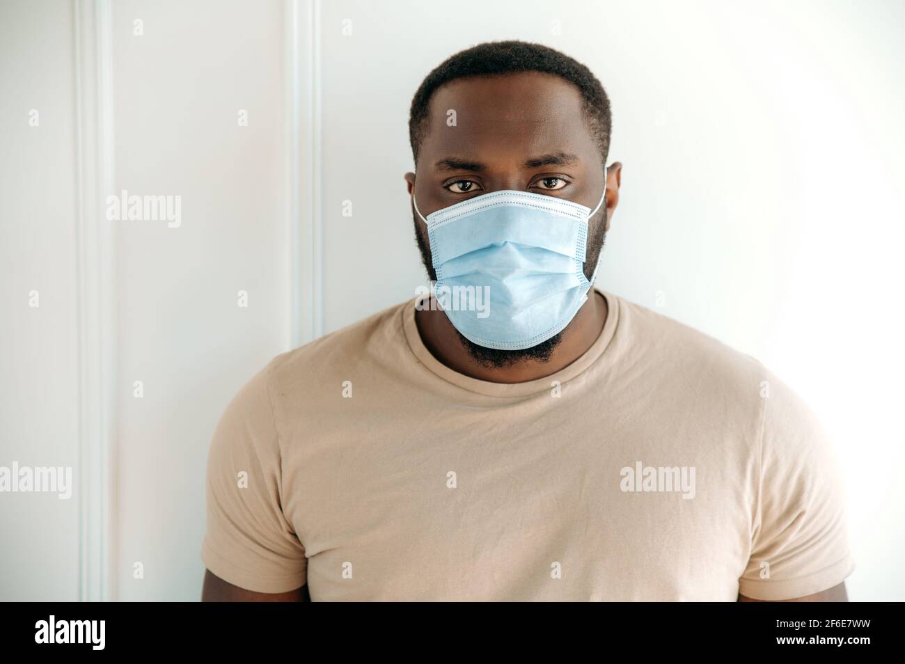 Précautions à mettre en quarantaine. Homme afro-américain, en tenue décontractée debout à l'intérieur avec masque médical de protection sur son visage et regardant directement dans l'appareil photo, rester à la maison Banque D'Images