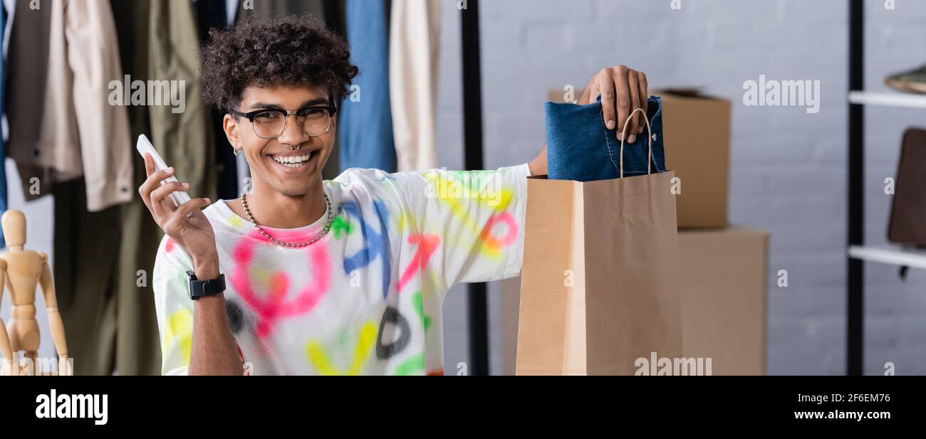 Joyeux propriétaire de petite entreprise afro-américaine avec un smartphone mettant des vêtements dans un sac de shopping, une bannière Banque D'Images