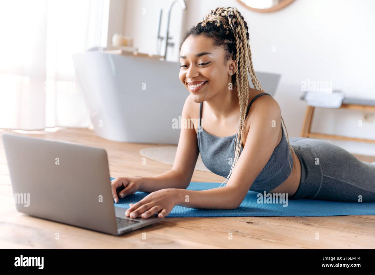 Athlétique belle jeune femme afro-américaine avec une bonne figure, se trouve sur le sol à la maison, montres en ligne cours de fitness utilise ordinateur portable, va dans pour le sport, mène un style de vie sain, sourire Banque D'Images