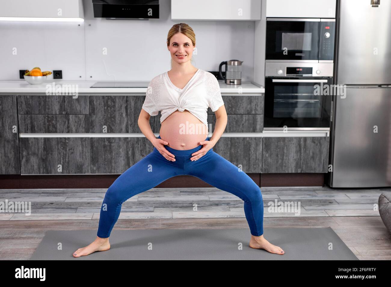 portrait de la femme faisant des exercices de yoga, debout avec les jambes écartées et souriant. dans la cuisine à la maison, grossesse, concept de mode de vie sain Banque D'Images