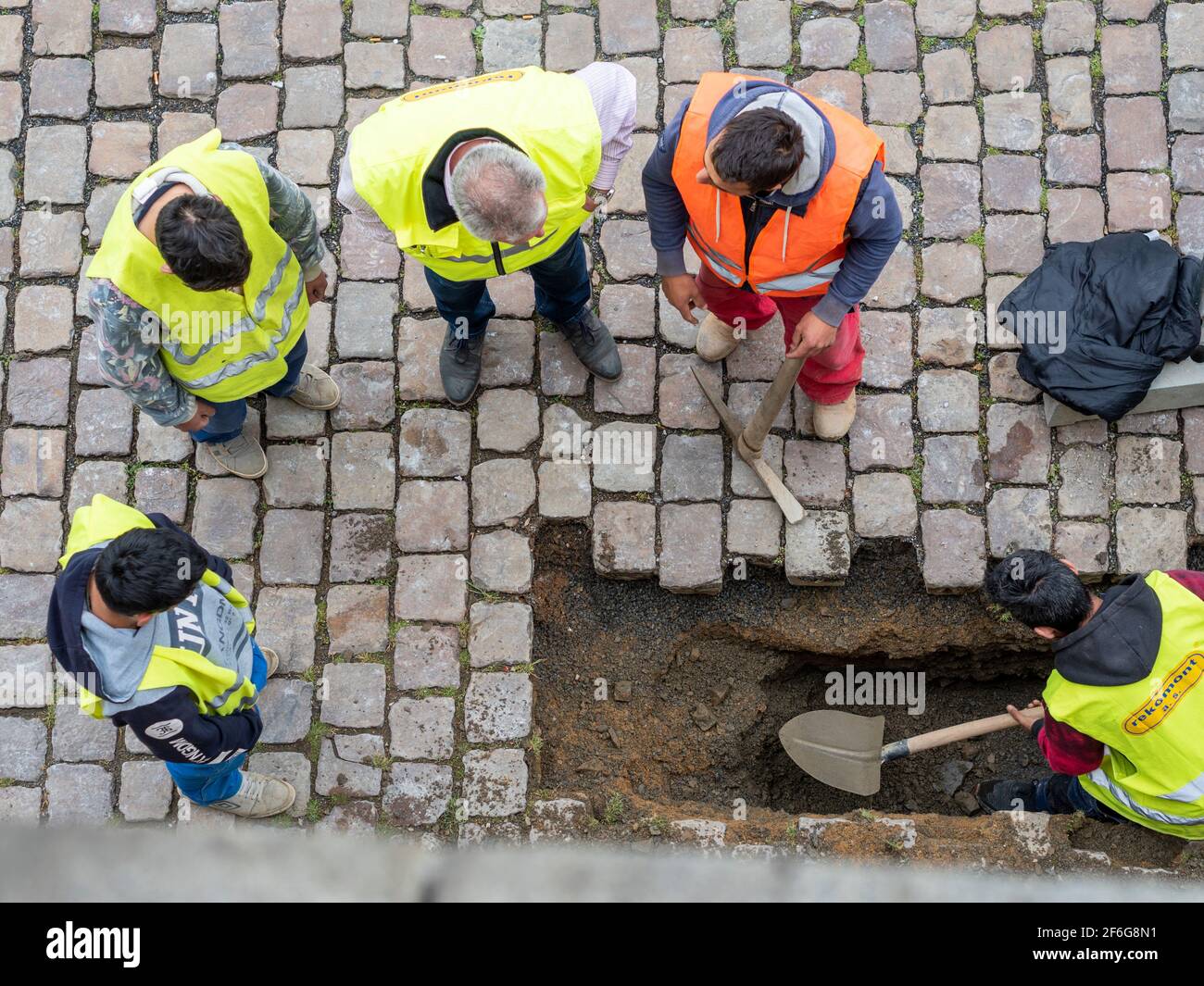 Supervision majeure des travaux de rue : un travailleur creuse un trou tandis qu'un autre tient un choix en parlant à un superviseur. Deux autres regardent. Banque D'Images