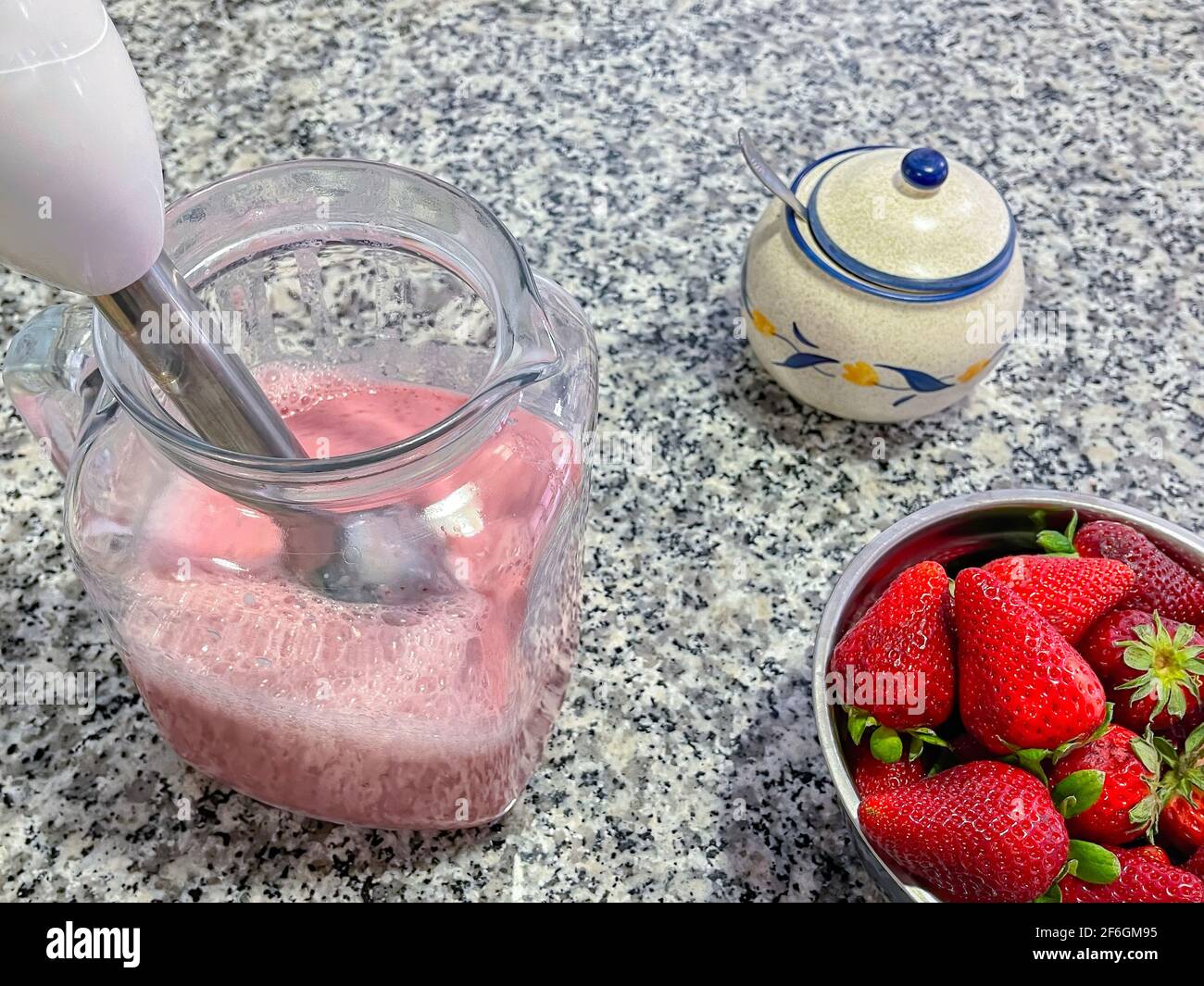 bol rempli de fraises et un pichet avec un milkshake aux fraises fraîchement préparé, un bol à sucre avec une cuillère à l'intérieur, un mixeur plongeant dans le bol, ingrédients Banque D'Images