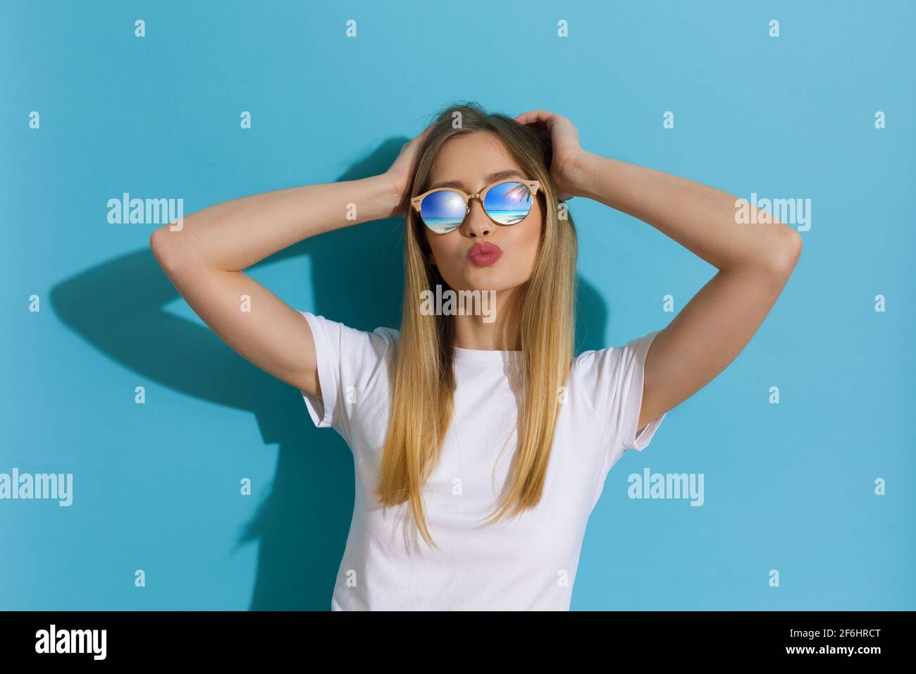 Jeune femme blonde en chemise blanche et lunettes de soleil pose avec la tête dans les mains et envoie un baiser. Taille relevée sur fond bleu. Banque D'Images