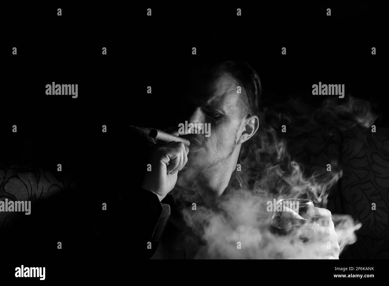 Homme fumant un cigare avec un verre de whisky dans le Main sur un arrière-plan noir foncé isolé.photo créative en noir et blanc portrait avec un nuage de Banque D'Images