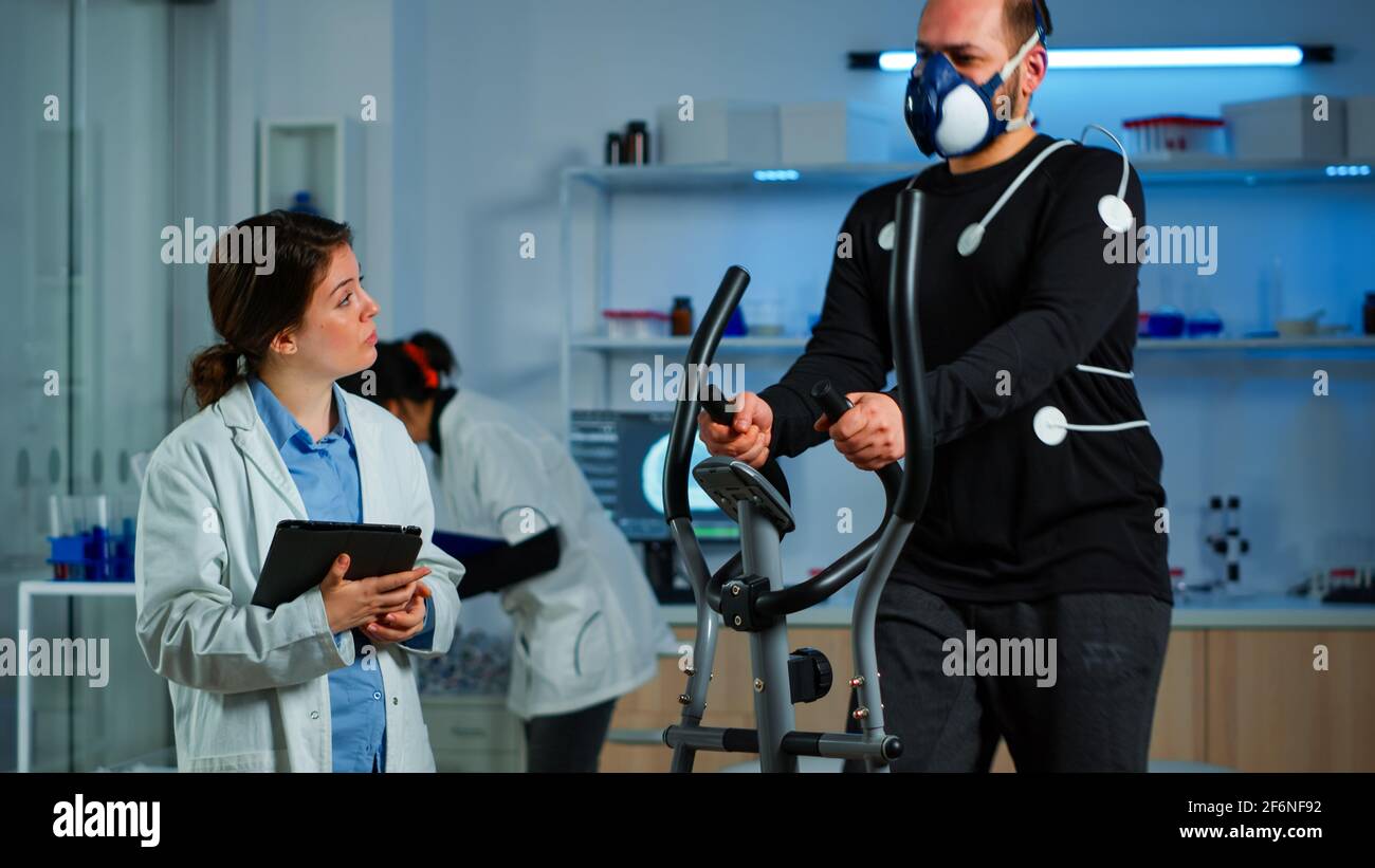 Chercheur médical discutant avec un sportif tout en mesurant son endurance  à l'aide de capteurs, d'électrodes et de masque de surveillance du rythme  cardiaque. Patient en train de courir en cross trainer