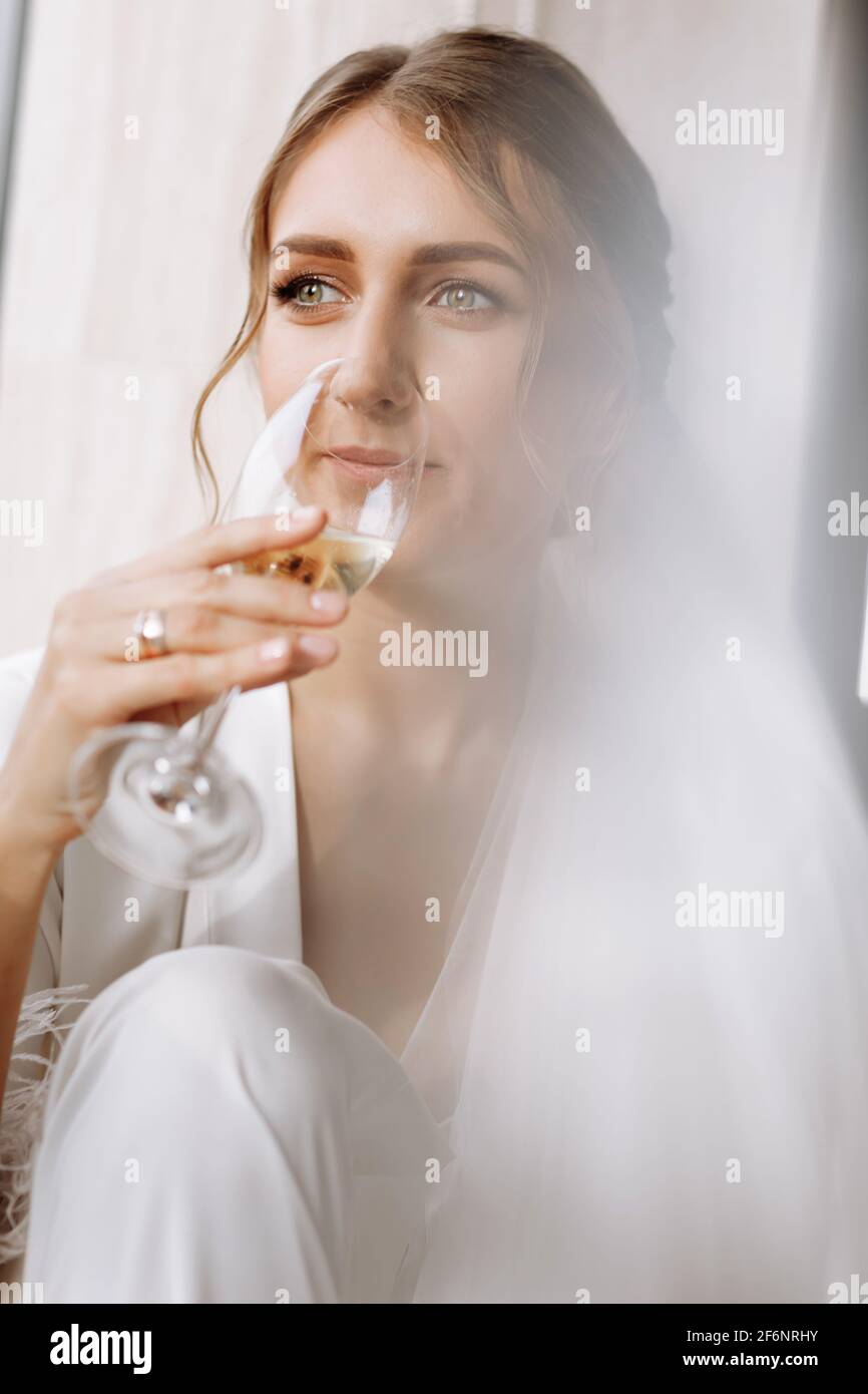 Le matin de la mariée. La mariée boit du champagne dans le peignoir. La jeune femme est assise sur une grande fenêtre dans une chambre d'hôtel dans la salle de bains. Belle fille en whi Banque D'Images