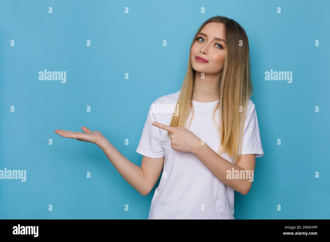 Décontracté jeune femme en chemise blanche tient la main levée, pointant et montrant quelque chose. Taille haute studio tourné sur fond bleu. Banque D'Images